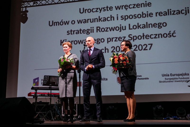 Trzech przedstawicieli LGD dziękuje za współpracę z Marszałkiem. Przemawia mężczyzna. Dwie kobiety trzymają kwiaty.