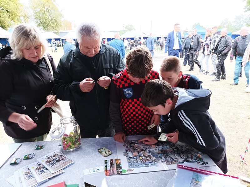 Po prawej trzech chłopców gra w grę "Tropienie zwierząt". Po lewej przy stole stoją dorośli  czytają ulotki.