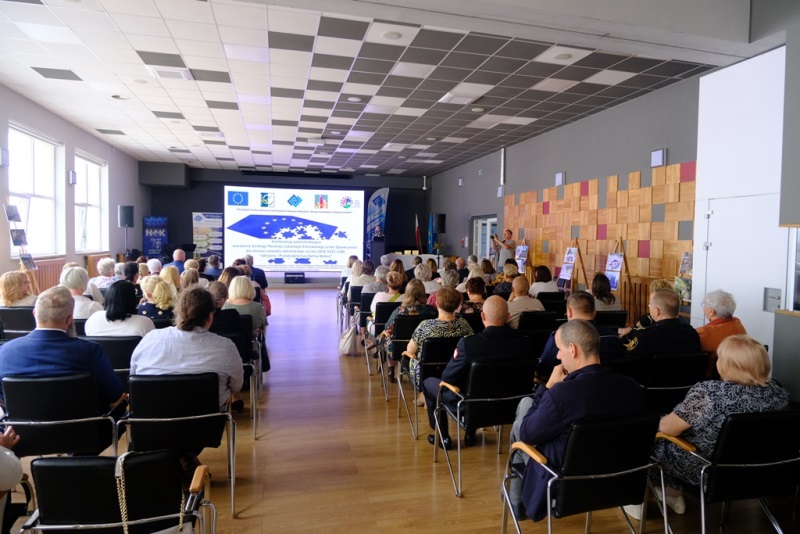 Uczestnicy konferencji siedzą na krzesłach (blisko 70 osób) i słuchają stojącego za mównicą prelegenta.