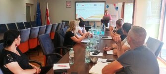 Uczestnicy szkolenia na pierwszym planie - siedzą przy stole i słuchają prelegenta, którego widać w tle.