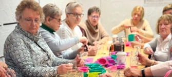 Seniorki siedzą przy stole i wykonują prace plastyczne.